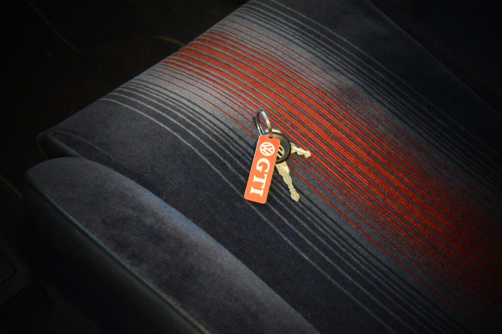 A set of keys with a red and white 'GTI' keychain lies on a car seat with a distinctive dark fabric pattern with red and blue stripes