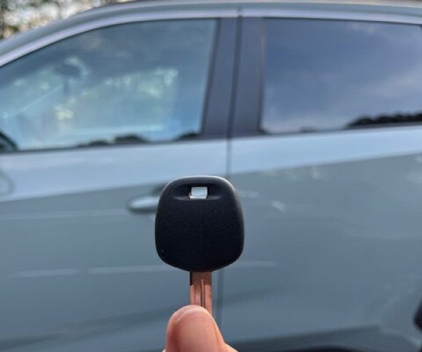 a person's hand holding a car key with a black fob in the foreground with a slightly different angle, focusing on a parked car and surrounding trees, all blurred in the background.