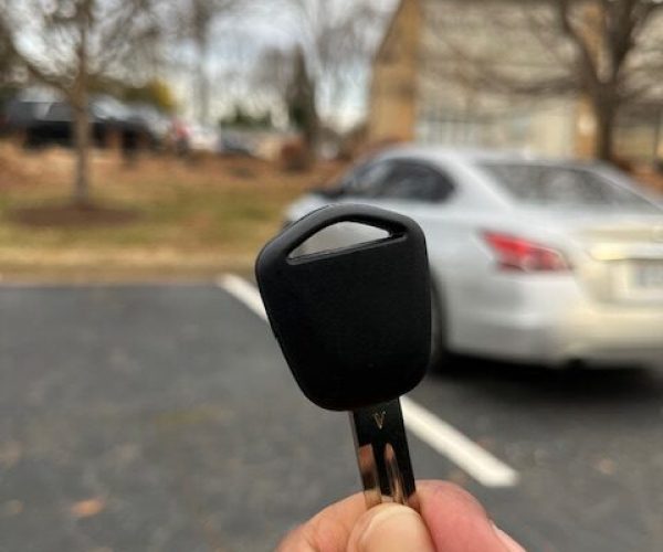 A photo focused on a car key in the foreground with a partial view of a white car's side and rear window blurred in the background.